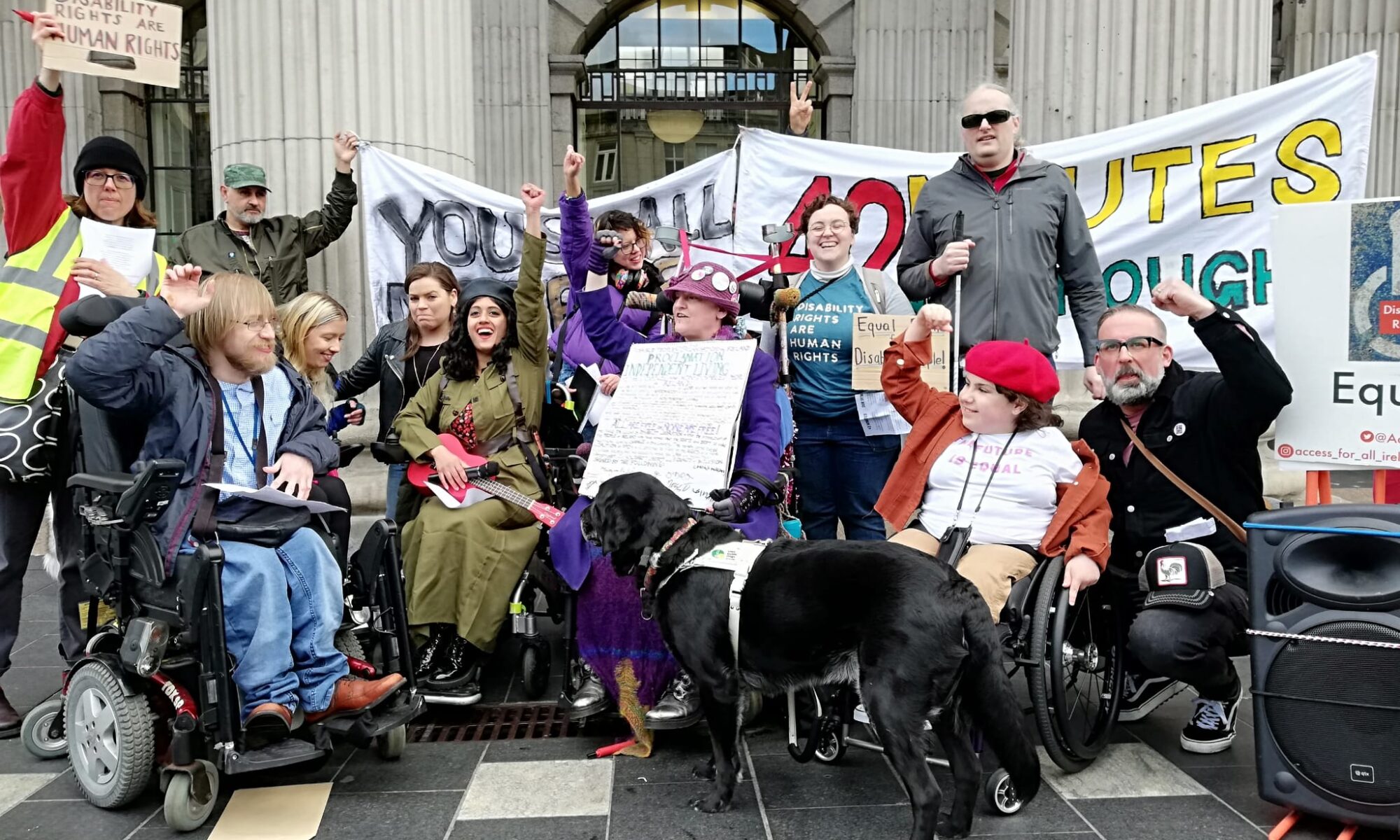 Image shows a group of protesters with raised fists outside the GPO. Included from the right are Ross Coleman, a man with blonde hair waving in a leather jacket and wheelchair beside him, Leesa Flynn with blonde hair smiling, Maryam Madani holding a red ukulele with raised fist and smile, with brown skin, black hair, black leather beret and Countess Markievicz costume, Isolde O'Brolcháin Carmody holding up Peadar's Declaration of Independent Living signed by attendees wearing a purple hat and sunglasses with her black lab assistance dog, Sophia Mulvaney wearing a red beret with raised fist on a wheelchair with white t-shirt and a pink-red cardigan, Bernard Mulvaney beside her wearing a black jacket with raised fist, a woman with long white hair with her golden lab assistance dog beside him and a sign which says "Equality". Behind them is Angela Mazzocco wearing a blue t-shirt, Declan Meenagh with his cane, Grainne Hallahan in a leather jacket, Charlie Mullowney smiling & protesters from the IRC campout