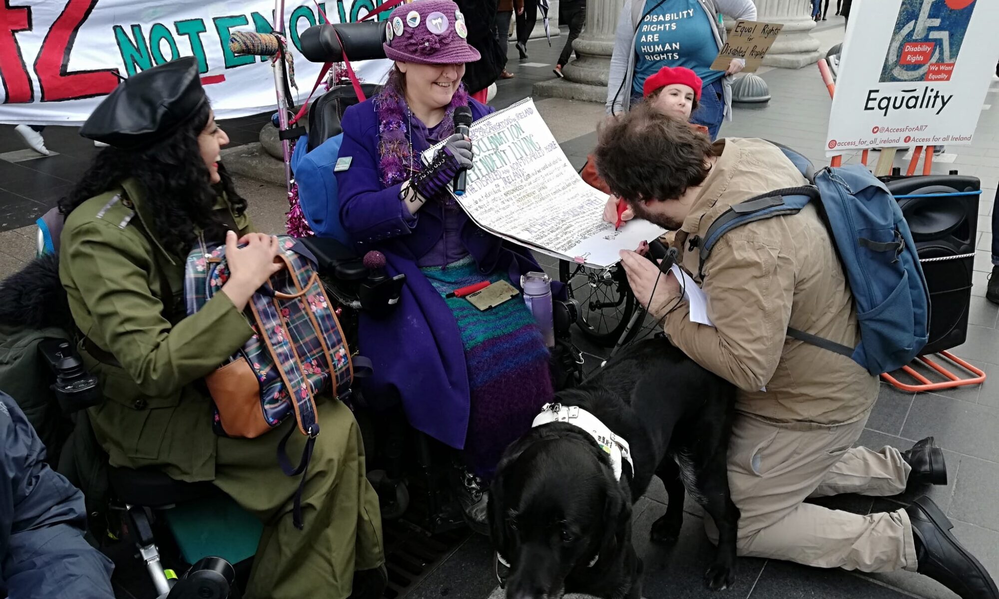 Image shows Peadar O'Dea kneeling before the Proclamation held up by Isolde. He is signing it. Isolde wears a 1916-inspired purple hat and outfit, is in an electric chair with her black lab assistance dog also at her feet. Peadar has dark curly hair and wearing khaki. Beside them is Maryam Madani with black curly hair and a black pleather beret, brown skin, wearing a Countess Markievicz-inspired outfit. Behind them is the banner "42 minutes is not enough". They are in front of the GPO, with other protesters around them.